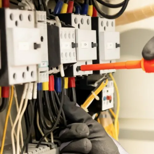 Electrician wearing gloves using a screwdriver to work on a complex electrical panel with multiple wires and circuits.