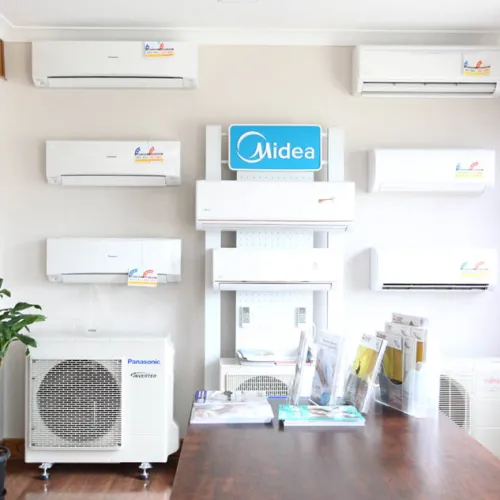 Wall-mounted air conditioners displayed in a showroom with a wooden table and potted plant indoor.