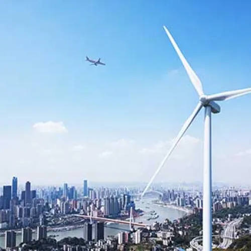 Modern city skyline with river, bridge, airplane in sky, and a large white wind turbine in the foreground under a clear blue sky