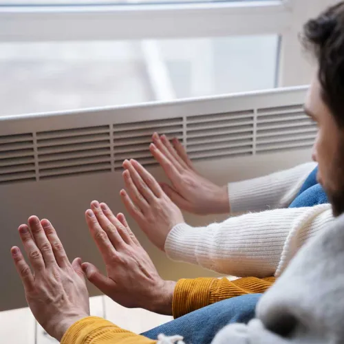Two people warming their hands near a heater by a window on a cold day indoors.