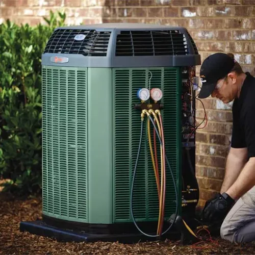 Technician repairing a green outdoor air conditioning unit beside a brick wall and shrubs during daytime