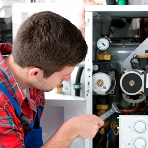 Technician in plaid shirt and blue overalls repairing a boiler with tools in a mechanical room.