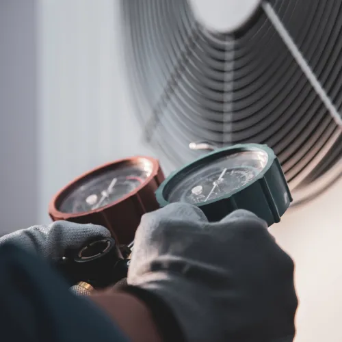 Technician wearing gloves adjusting HVAC manifold gauges near an air conditioner fan for system diagnostics.