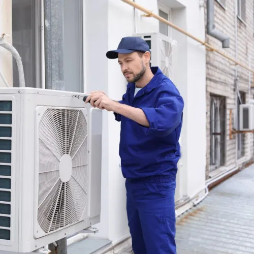 Technician in blue uniform repairing outdoor air conditioning unit on building exterior wall.