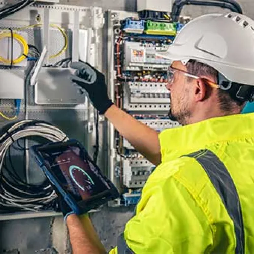 Electrician in safety gear inspecting circuit breaker panel using tablet for diagnostics and repairs