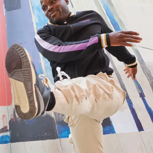 Man in black jacket and beige pants kicking leg forward against colorful urban wall background.