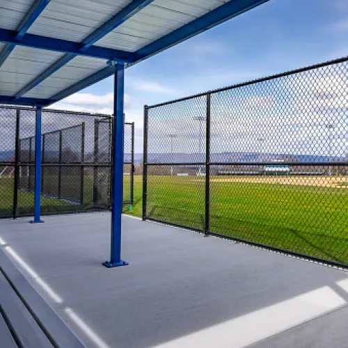 Empty baseball dugout with blue roof overlooking a green baseball field under a clear sky.
