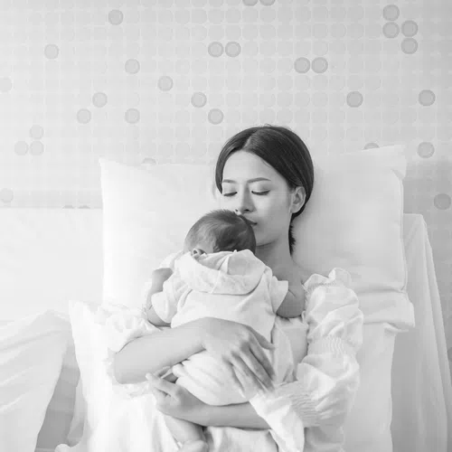 Mother resting on bed holding and comforting her baby in a calm, cozy bedroom setting.