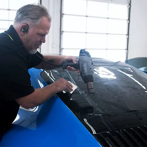 Man using heat gun to apply tinted film on blue car window in bright garage