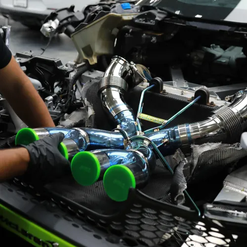 Mechanic installing shiny chrome turbocharger piping with green caps in a high-performance car engine bay.