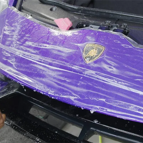 Person applying protective film on purple Lamborghini front bumper for paint protection.