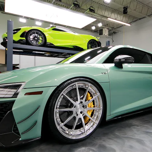 Matte green sports car with silver wheels and yellow brakes inside a modern garage with other luxury cars.