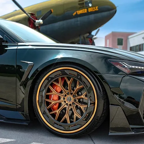 Close-up of a luxury car front wheel with gold rims and orange brake calipers parked near a vintage airplane.
