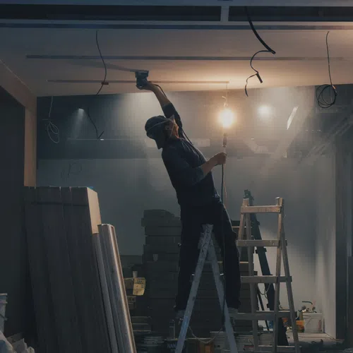 Worker wearing a mask standing on a ladder installing ceiling panels in a dimly lit construction site.