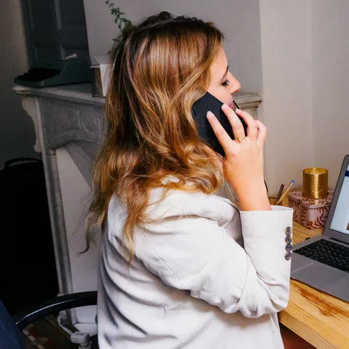 Woman in white blazer talking on phone while working on laptop at wooden desk indoors