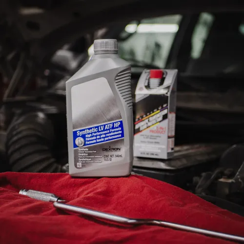 Bottle of Synthetic LV ATF HP automotive fluid on a red cloth with a wrench near a car engine.