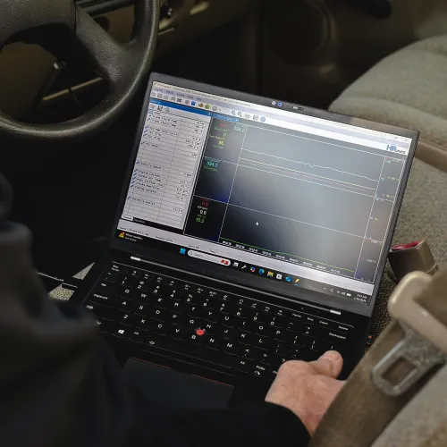 Technician using a laptop inside a car to diagnose vehicle performance data on screen.