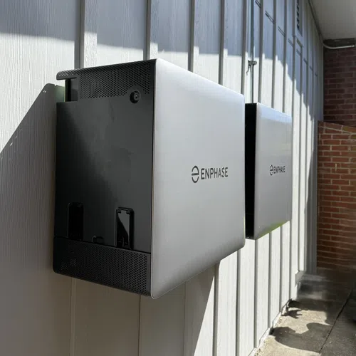 Two Enphase solar energy storage units mounted on a white wooden exterior wall in sunlight.