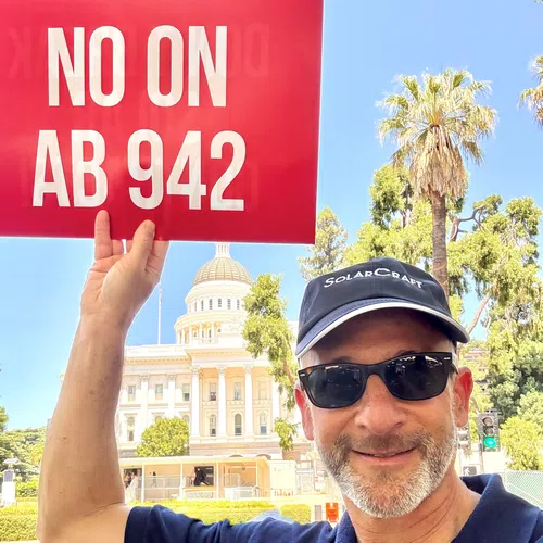 Man in sunglasses and SolarCraft cap holding a red sign saying NO ON AB 942 with a capitol building and palm trees in background
