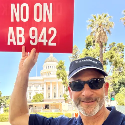 Man in sunglasses and SolarCraft cap holding a red sign saying NO ON AB 942 with a capitol building and palm trees in background