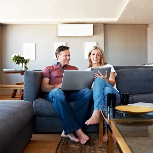 Couple sitting barefoot on gray couch using laptop with heat pump mini-split air conditioner on the wall