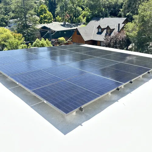 Array of solar panels installed on a white foam Eichler rooftop with green trees and houses in the background on a sunny day