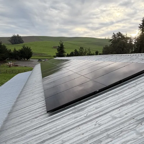 Solar panels installed on a corrugated metal roof with green hills and trees under a cloudy sky.