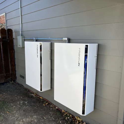 Two white Franklin energy storage batteries mounted on the exterior wall of a house next to a wooden fence.