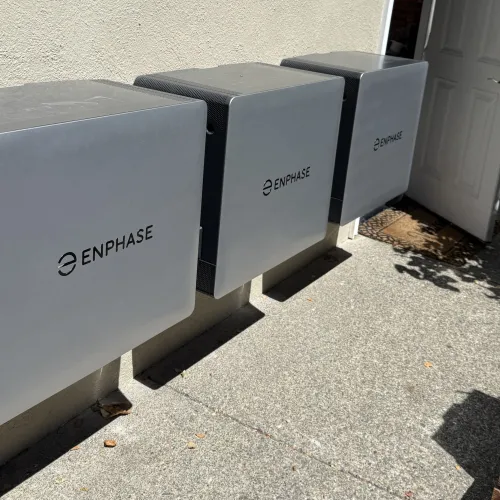 Three gray Enphase energy storage battery units mounted on an exterior wall near a white door on a sunny day.
