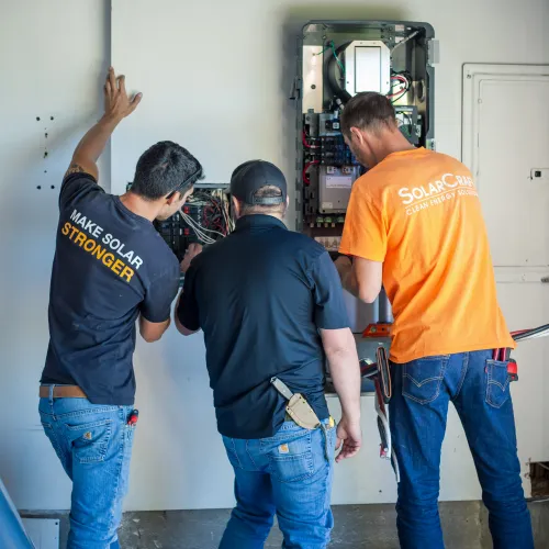 Three technicians working on electrical panels installing or repairing solar energy equipment indoors.
