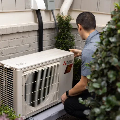 Technician inspecting a Mitsubishi Electric outdoor HVAC unit installed next to a house wall with surrounding greenery.