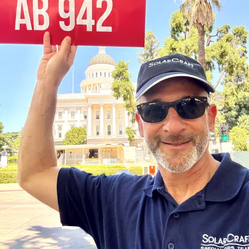Man holding a red sign saying NO ON AB 942 near a government building with palm trees on a sunny day