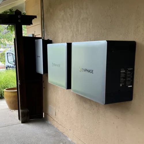 Enphase solar energy storage batteries mounted on an exterior beige wall near a wooden gate outdoors.