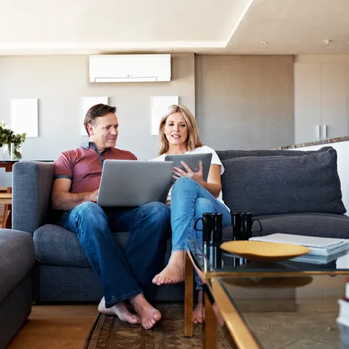Couple sitting on gray sofa using laptop and tablet in modern living room with natural light