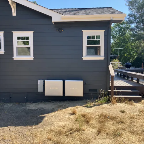 Dark gray house exterior with three white-trimmed windows, two enphase battery units, and wooden deck steps in dry yard.