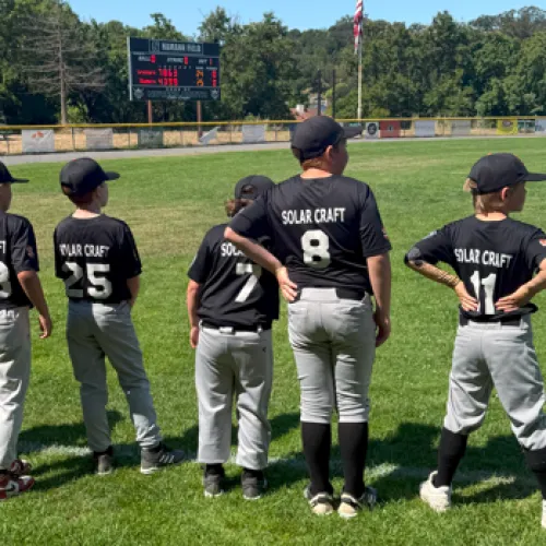 Youth baseball players in black Solar Craft jerseys stand on the field looking toward the scoreboard during a sunny day.