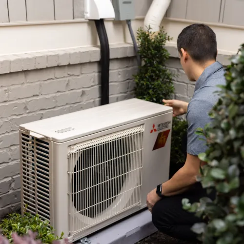 Technician inspecting Mitsubishi Electric outdoor air conditioning unit beside house exterior wall