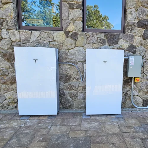 Two white Tesla Powerwall battery units installed on a stone wall exterior under windows with electrical wiring connections.