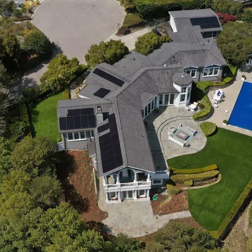 Aerial view of a modern large house with solar panels, swimming pool, patios, and manicured green lawn surrounded by trees.