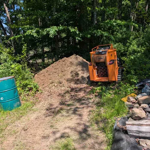 Orange SCAG skid steer loader next to a dirt pile in a forest clearing on a sunny day.