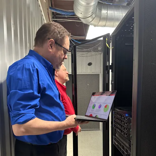 Two IT professionals in blue and red shirts working on server racks using a laptop with data charts.