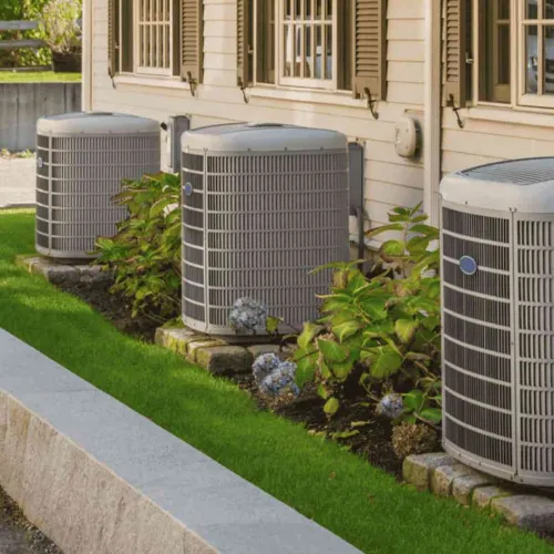 Three outdoor air conditioning units installed beside a house with beige siding and green plants.