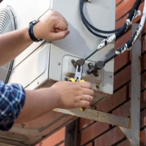 Technician repairing outdoor air conditioning unit with pliers on a brick wall-mounted bracket.