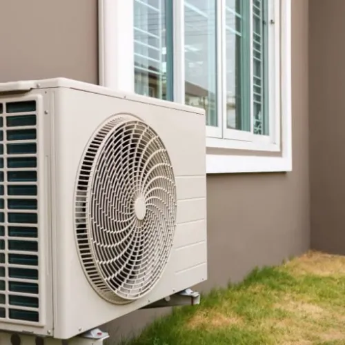 Outdoor residential air conditioning unit mounted on wall near grass and windows on beige house exterior.