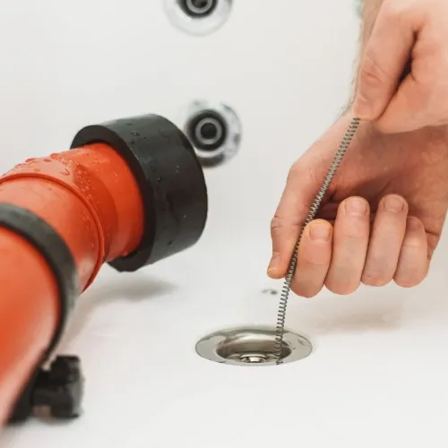 Person using a drain cleaning spring tool to clear a clogged bathtub drain with visible red pipe underneath.