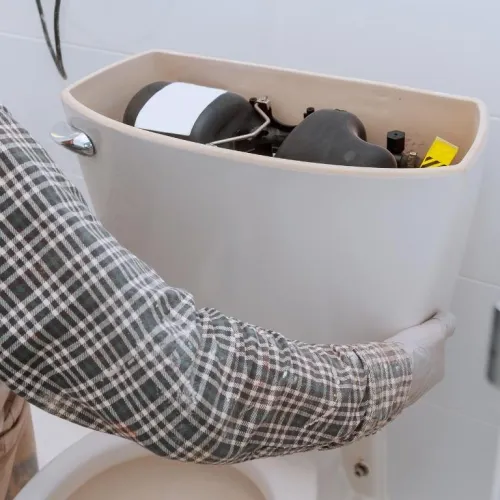 Person repairing a toilet tank, holding the tank lid and wearing a plaid shirt in a bathroom.