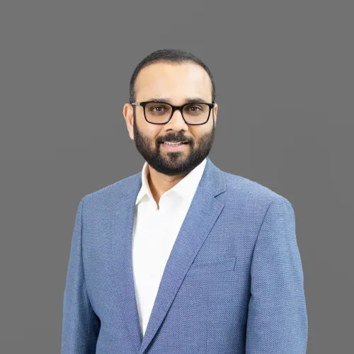 Bearded man with glasses in a blue blazer and white shirt smiling against a gray background