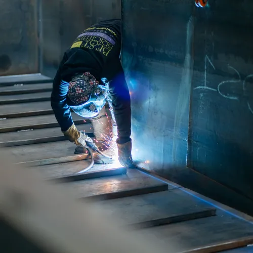 Welder using protective gear welding metal beams with sparks flying in an industrial workshop.