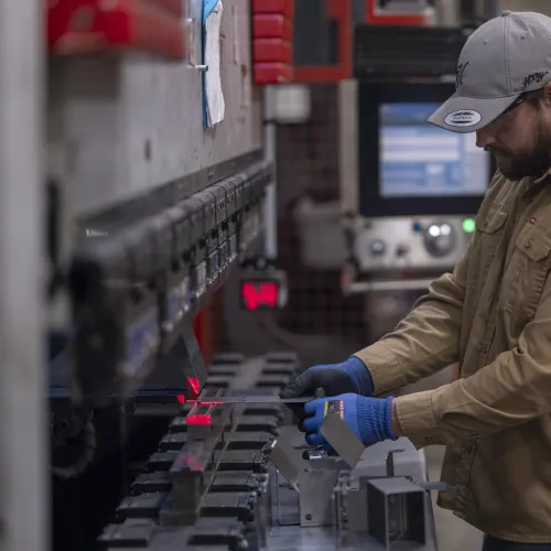 Worker in tan jacket and cap operating industrial machine with metal parts in a factory setting.