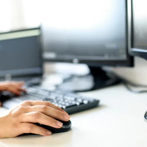 Person using computer mouse and keyboard with multiple monitors on a clean office desk workspace.